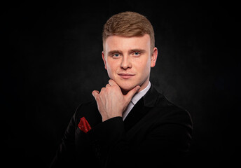 Portrait of a handsome young redhead man wearing a formal black suit and red pocket square, posing with his hand on his chin in a thoughtful and confident manner against a dark studio background.