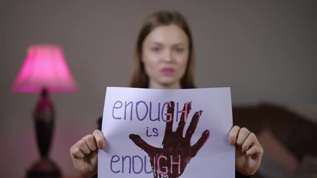 White woman holds enough is enough sign with red handprint, assertive expression in bedroom setting with pink lamp, closeup protest visual conveying outrage,