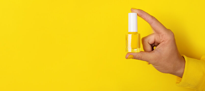Close-up of a hand in a yellow sleeve holding a clear nail polish bottle against a bright yellow background, minimalist beauty concept. - Powered by Adobe