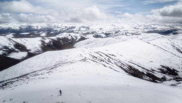 Aerial view of a lone figure stands amidst the stark white snow-covered peaks, contrasting with the dark, rugged terrain beneath a cloudy sky, Wenshan Zhuang and Miao Autonomous Prefecture, China.