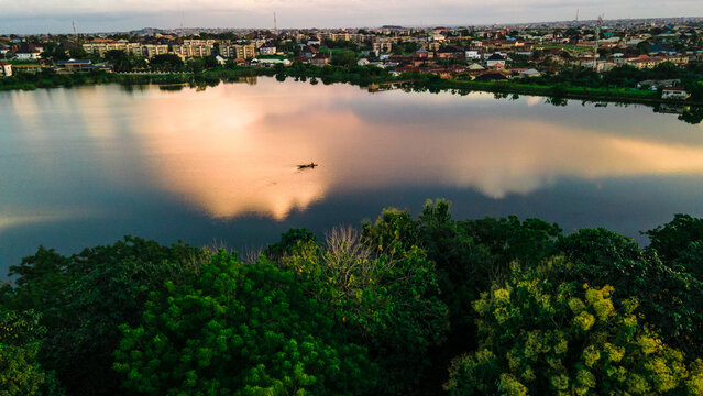 Aerial view of the tranquil lake mirroring the soft glow of the sky, embraced by lush trees, with buildings visible in the background, Ilorin, Kwara, Nigeria.
