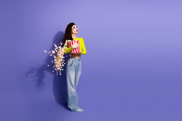 Young woman in neon yellow top stands against purple background holding popcorn bucket while wearing 3d glasses for fun fashion and shopping vibe