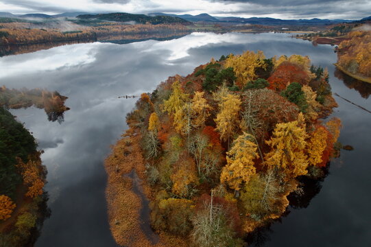Aerial view of an island ablaze with autumnal hues amidst the tranquil, reflective waters under a brooding sky, Kingussie, Scotland, United Kingdom.