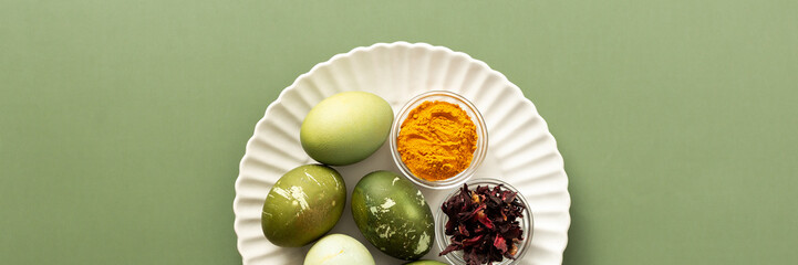 A decorative plate holds several dyed Easter eggs, showcasing shades of green, alongside bowls containing turmeric and dried flowers hibiscus for natural coloring, how to dye eggs marble green, banner