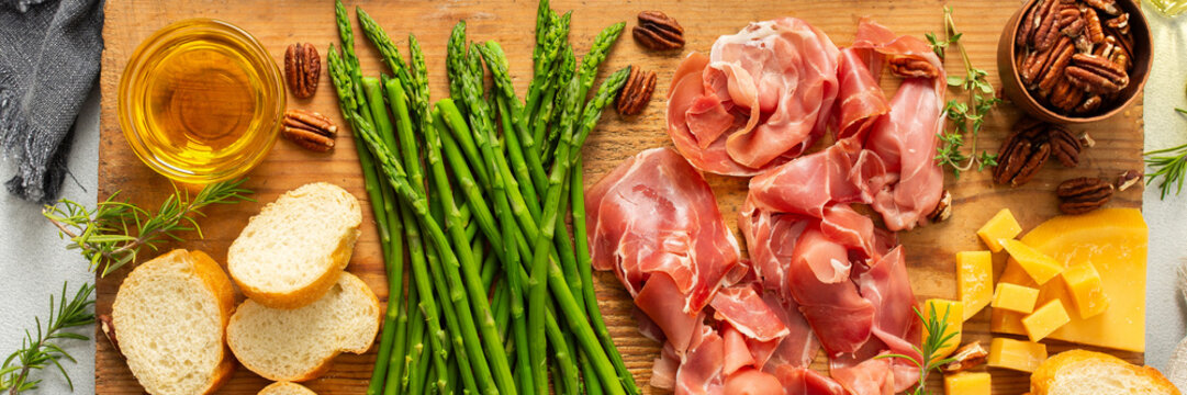 A beautifully arranged charcuterie board features fresh asparagus, various meats, cheeses, and nuts, set on a wooden plank with slices of bread. Perfect for sharing at gatherings, banner