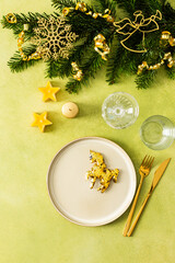 A festive Christmas table setting with an empty plate and cutlery on a bright green background. Spruse branches and golden tree decorations create a warm, holiday atmosphere