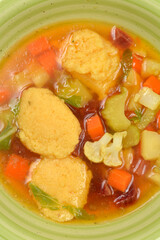A vertical close-up shot focuses on a rich yellow broth filled with semolina dumplings and chunky diced vegetables, including carrots and cauliflower, in a green bowl