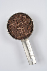 A stainless steel one-third cup measuring cup filled with coarsely shaved dark chocolate is centered on a solid white background, viewed from above