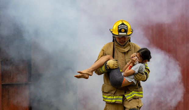 Firefighter in fireproof clothe is carrying young kid out of the burning building full of ash and smoke for fire extinguish risk rescue mission and accident management usage