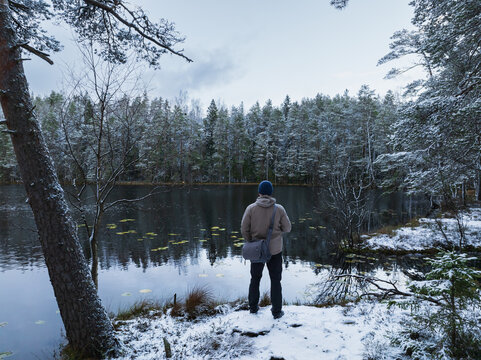 Male hiker standing on the snowy bank of a dark forest lake in an Estonian bog during winter, aerial drone view.