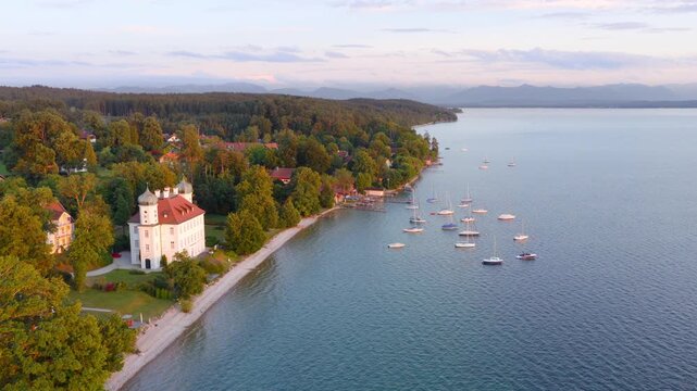 Soft evening light rests over Lake Starnberg. Quiet boats float on the clear water, while the shoreline and its distinctive building glow warmly in the fading daylight.