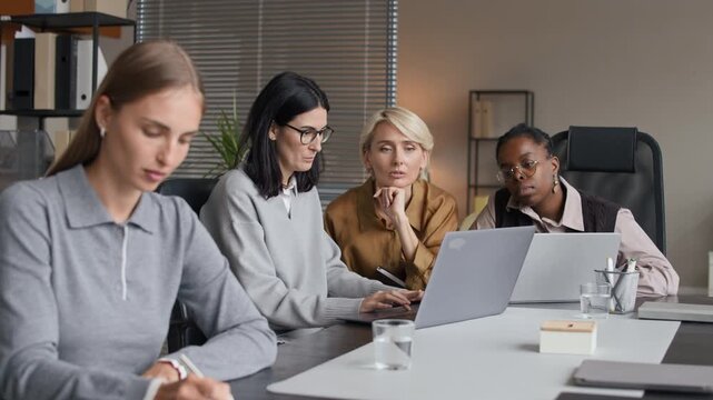 Rack focus shot of concentrated young Caucasian female employee drafting project on paper with her multiethnic female office colleagues brainstorming in front of laptops in background