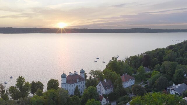 Evening light over Lake Starnberg. The sun sets behind the horizon while a castle and houses sit along the shore surrounded by dense green forest.