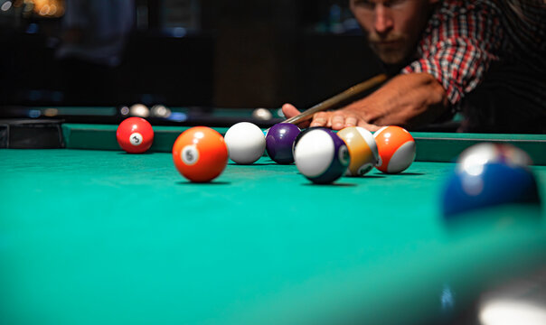 A close-up view of colorful billiard balls scattered on a green felt table while a focused man in a plaid shirt aims his cue stick in the blurred background.