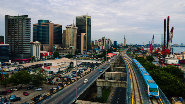 Aerial view of a modern blue train traversing along the tracks against the backdrop of towering skyscrapers and bustling city life, Lagos, Nigeria.