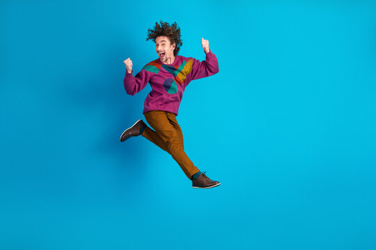 Young man jumping with joy against blue backdrop wearing a colorful sweater brown pants and stylish shoes conveying energy fun and positivity