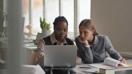 Medium shot of young multiethnic female colleagues sitting at desk in modern well-equipped open space office and working on business project together, analyzing paper report and data on laptop - Powered by Adobe