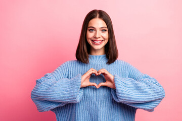 Young woman in blue knit sweater making heart shape with hands against pink background