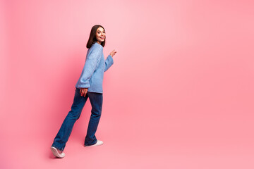 Fototapeta premium Young woman in blue sweater walks on pink background smiling and looking back