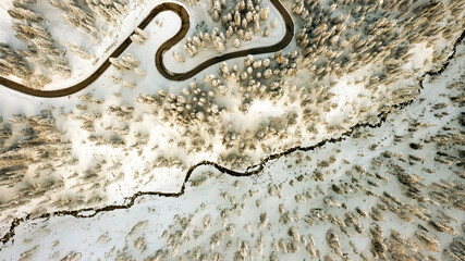 Aerial view of a winding road cutting through a snow-laden forest alongside a dark, meandering stream, Passua, Trentino-Alto Adige, Italy.