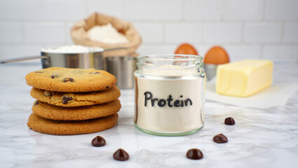 Protein powder in glass jar with chocolate chip cookies and baking ingredients on marble kitchen counter,  healthy baking, dietary supplement, glp-1 meal, glp-1 food, glp-1 recipes, high protein food