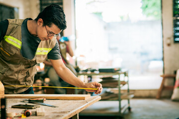 Young cabinetmaker measuring a piece with a tape measure in the workshop. Carpenter with glasses taking measurements at the workbench