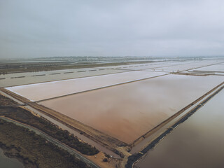 Aerial view of salt marshes reflecting the cloudy sky, creating a muted palette of pinks and grays in the Bacuta Salt Marshes, Huelva, Andalucía, Spain.