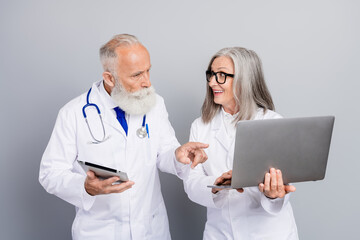 Two aged doctors discuss patient care using tablet and laptop in a clinic setting with stethoscope and glasses