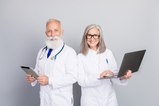 Two doctors aged and experienced colleagues in white coats stand in a clinic using tablet and laptop for medical consultation and patient care