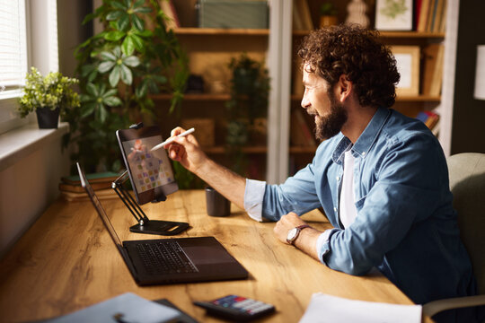 A man engages in focused work at his home office, using a digital tablet and laptop on a wooden desk, surrounded by plants and natural light streaming through a window.