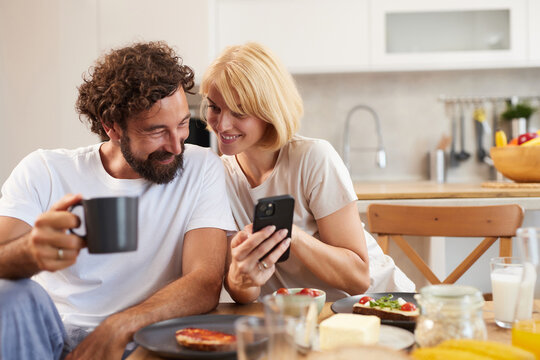 A couple is seated at their kitchen table, smiling and sharing a joyful moment. They are enjoying brunch with coffee, breakfast dishes, and engaging with a smartphone together.