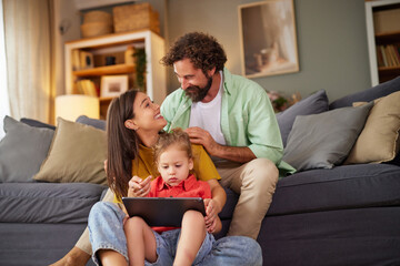 A family spends enjoyable time together in their living room. The parents are engaged with their children, sharing smiles while one child plays with a tablet.