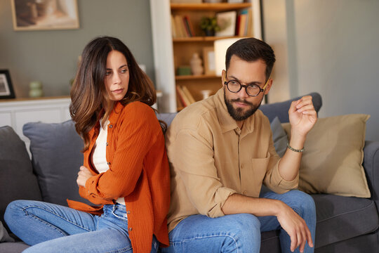Two individuals are sitting on a sofa in a cozy living room, showing signs of tension in their relationship.