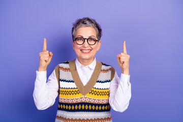 A mature woman with short gray hair and glasses points up with both fingers wearing a colorful...