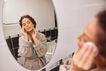 A woman with curly hair is gently applying skincare to her face while looking into a round mirror. She is wearing a cozy robe in a bright and modern bathroom setting.