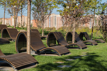 Modern, comfortable wooden benches with wave-like shape are located in green park against backdrop of trees and city buildings. Cloud Park, Krasnodar Public Park, or Galitsky City Park.