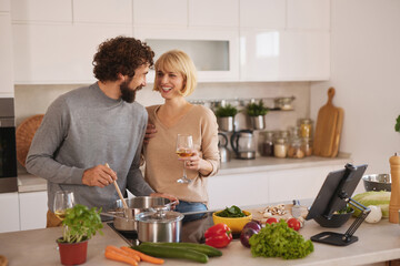 A couple is happily cooking together in a stylish kitchen. They are smiling at each other while preparing a meal and enjoying a drink. Fresh ingredients are all around.