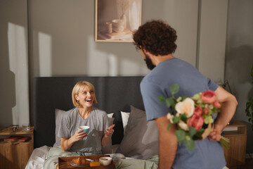 A woman smiles as she enjoys her breakfast in bed. A man stands nearby, hiding a bouquet of flowers behind his back. It is a warm morning in a comfortable bedroom.