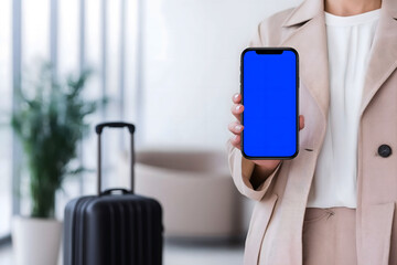 Businesswoman in a stylish beige suit holds smartphone with blue screen, standing in modern lobby with suitcase and greenery, showcasing professional travel readiness and technology