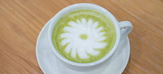 A warm cup of matcha latte rests on a white saucer atop a wooden table, with white foam forming a simple floral design in the center.