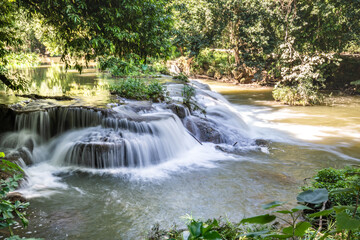 Obraz premium A small waterfall cascades over layered rocks surrounded by trees, with the water flowing in shallow steps into a wider pool.