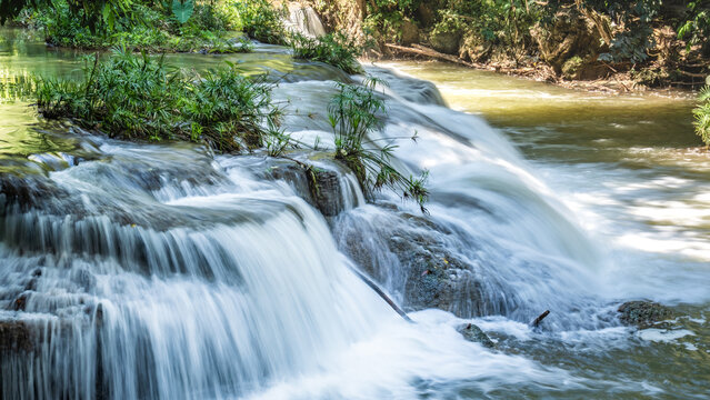 A small waterfall cascades over layered rocks surrounded by trees, with the water flowing in shallow steps into a wider pool.