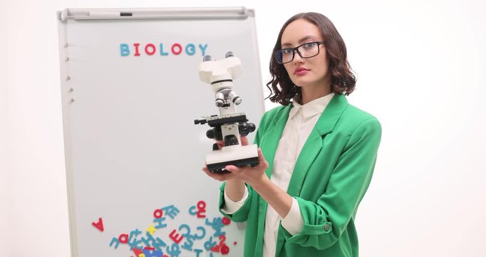 Female teacher takes off glasses to look in microscope at biology lesson against whiteboard. Woman scientist studies biomaterial specimen in classroom