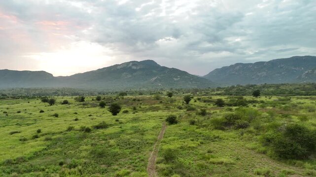 Wide drone panorama near Benguela, Angola, showing wild grasslands, distant mountains, and soft sunlight piercing through dramatic clouds at dusk