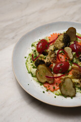 Close-up of an artisan vegetable salad featuring cherry tomatoes, pickled cucumbers, carrots, and broccoli, served with a creamy white dressing on a light marble table