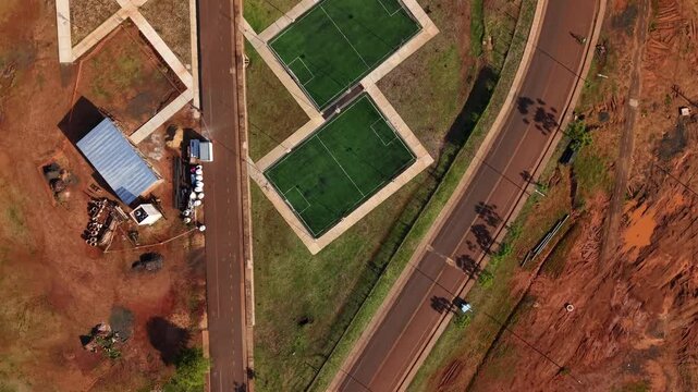 New five-a-side football soccer fields sit alongside a curved urban road and construction site, bright green synthetic pitches bordered by walkways, in Posadas, Misiones, Argentina.