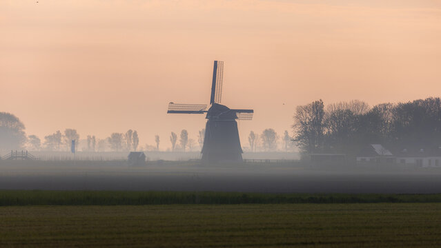 View of a solitary, silhouetted windmill stands majestically against a soft, hazy dawn sky, its dark form a striking contrast to the muted pastel colors, Schagen, North Holland, Netherlands.