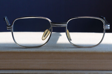  Stylish vintage reading glasses with clear lenses lie on stacked books in bright sunlight with light reflections on the lenses on a wooden table, dark background, close-up.