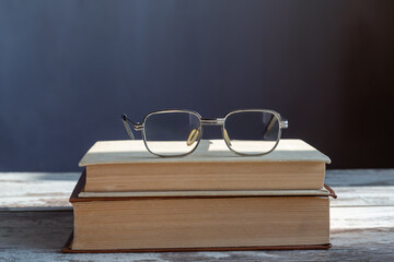 Glasses with clear lenses lie on two stacked books in bright sunlight on a wooden table, dark background, close-up.