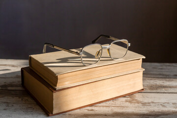 Stylish vintage reading glasses lie on stacked books in bright sunlight with light reflections on the lenses on a wooden table, dark background, close-up.
